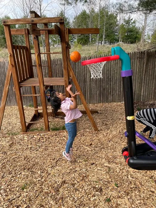A young child jumps to shoot a basketball into a toy hoop beside a wooden playset in a wood-chip yard