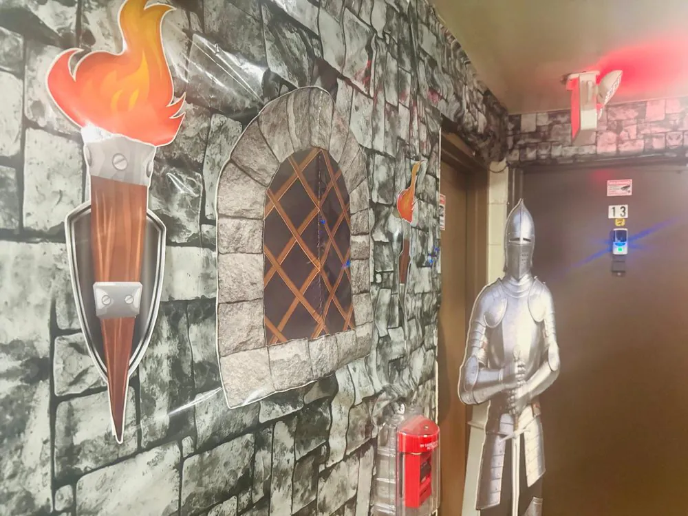EduCastle corridor with stone-wall murals, a painted torch, an arched window and a knight cutout by the door