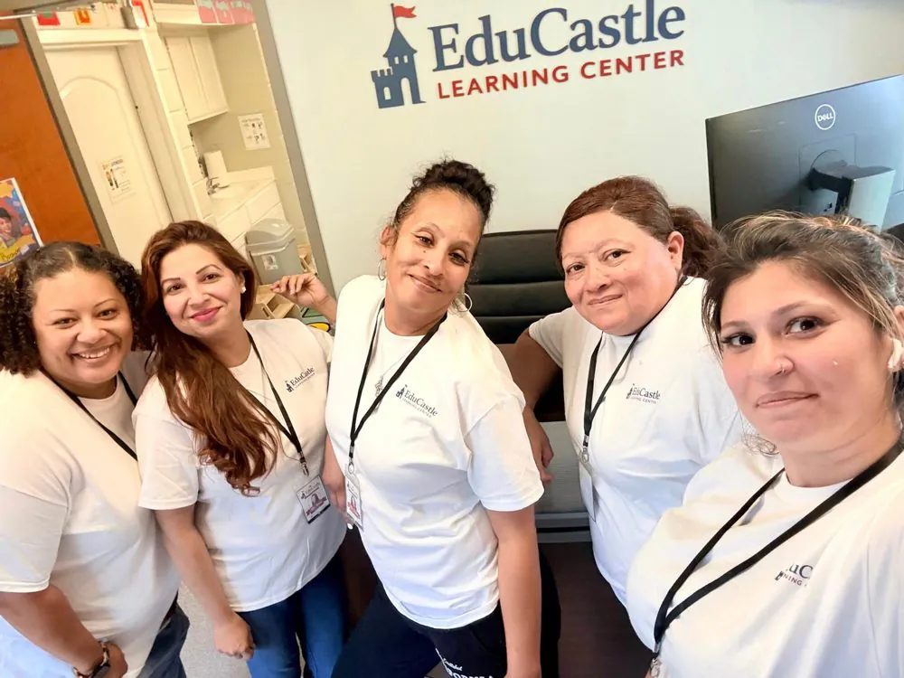 Five smiling EduCastle teachers in matching white shirts and lanyards at the front desk under the center&rsquo;s logo