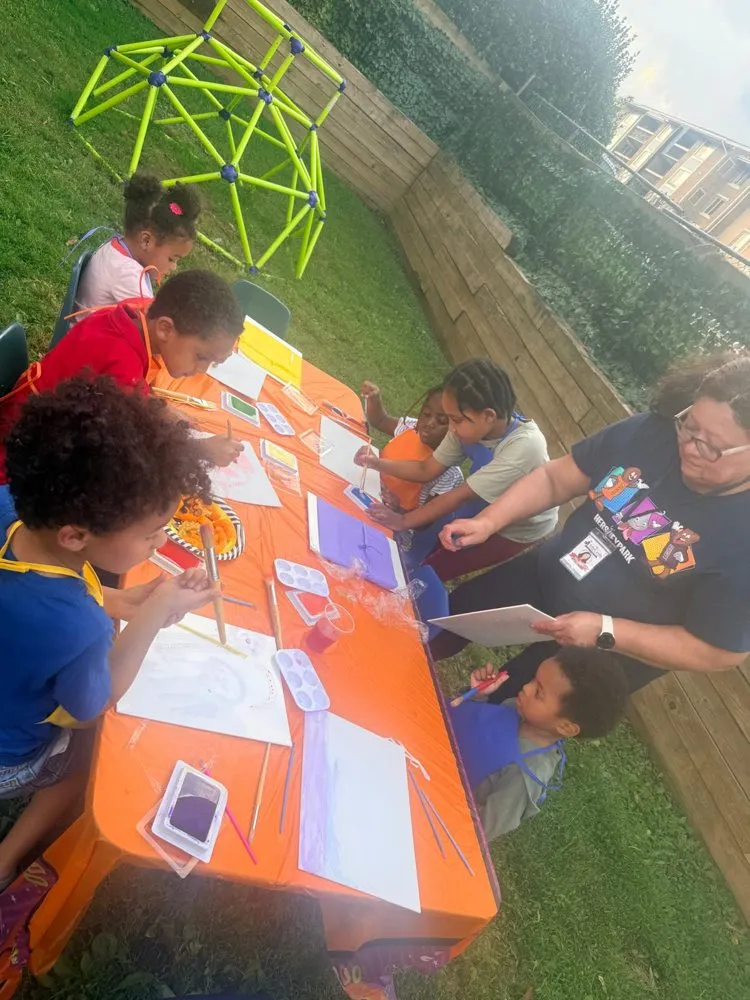 Children in aprons painting canvases at an orange table on the lawn with a teacher at the EduCastle fall festival
