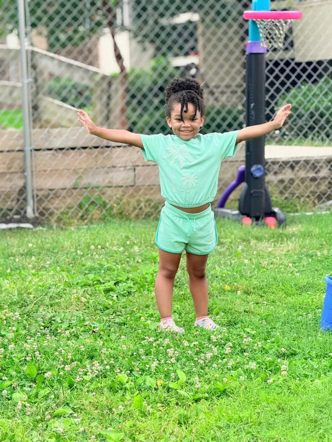 A smiling preschooler stands on the grass with arms stretched wide in the fenced EduCastle play yard