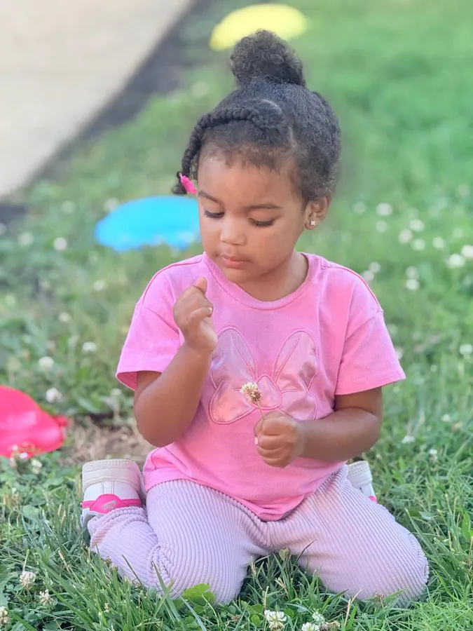 A toddler kneels in the grass and closely examines a small clover flower during outdoor time at EduCastle