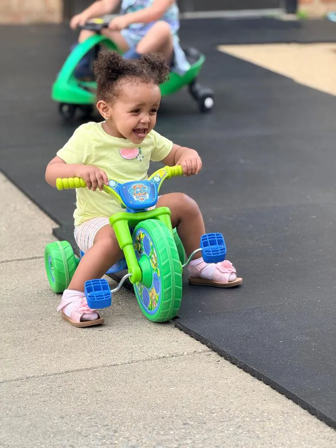 A laughing toddler pedals a green tricycle across the EduCastle playground while another child rides behind