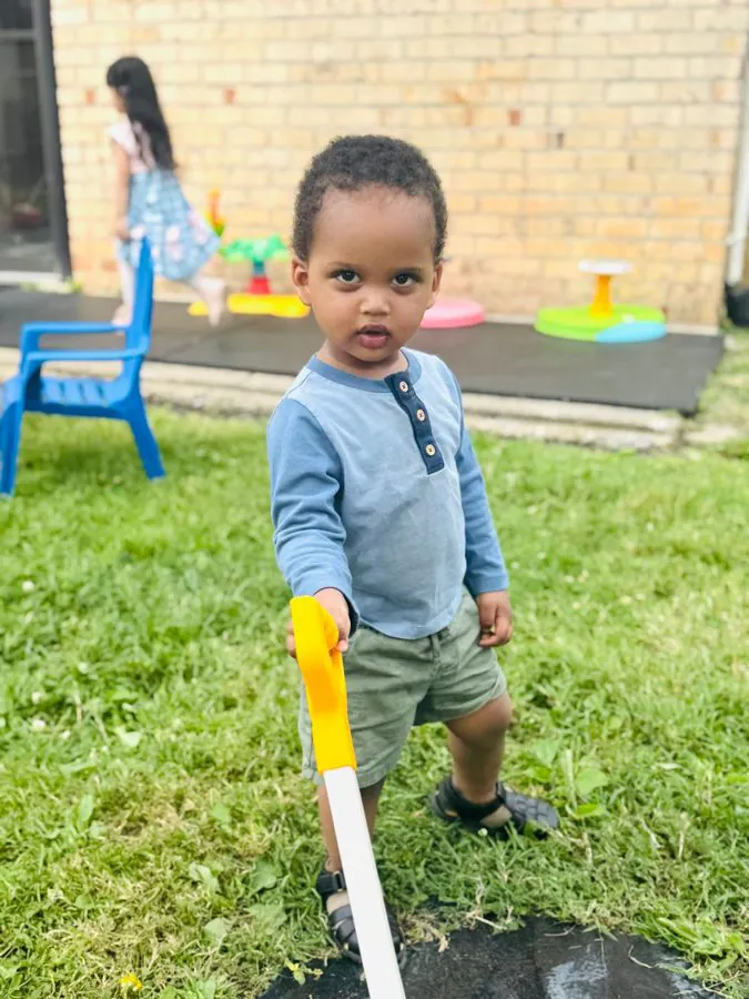 A toddler in a blue shirt pushes a toy yard tool across the grass in the EduCastle backyard play area