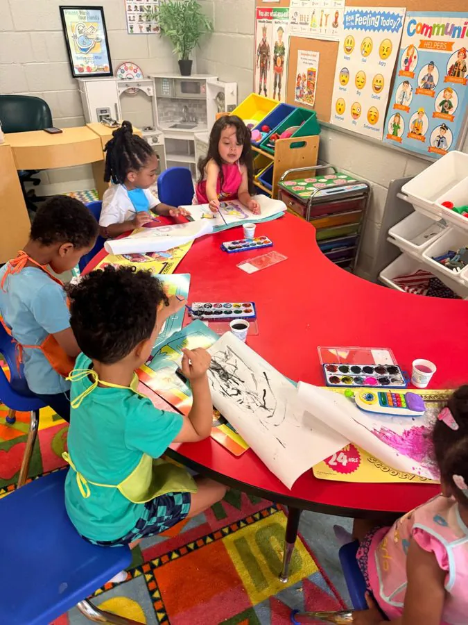Preschoolers in aprons paint with watercolors around a red table in an EduCastle classroom