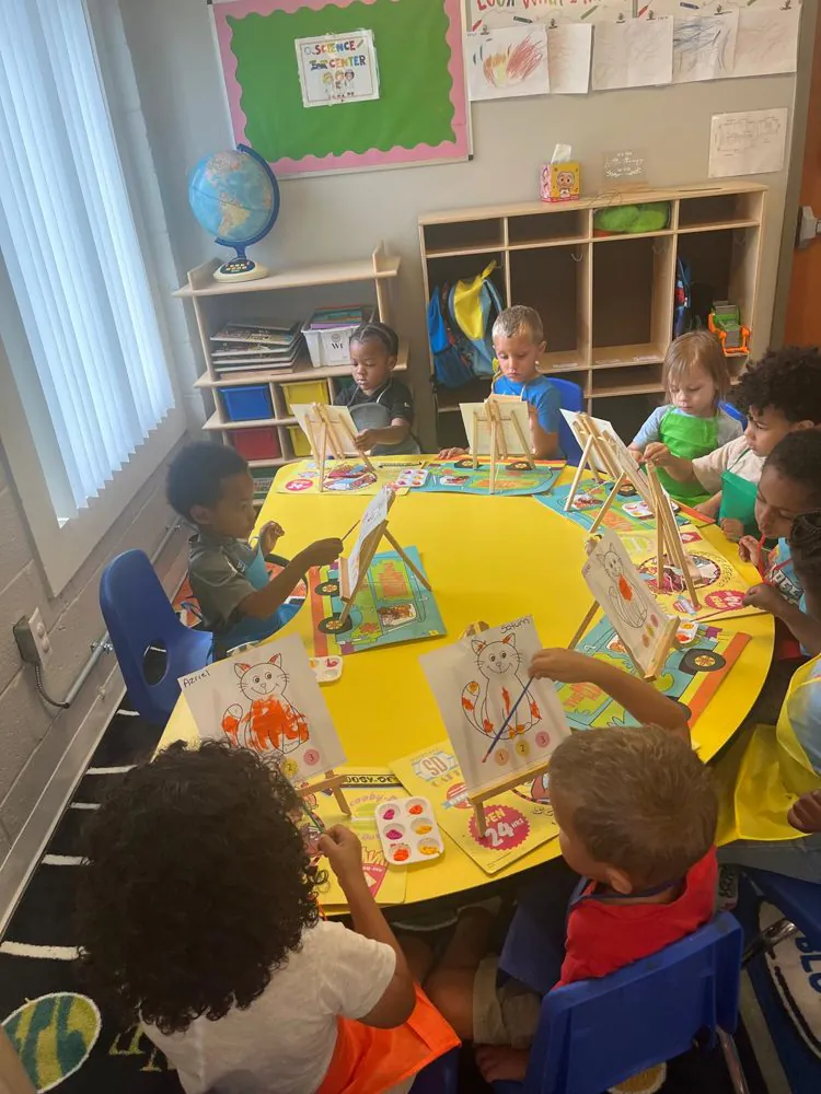 Preschoolers painting cats on small tabletop easels around a yellow table in an EduCastle classroom