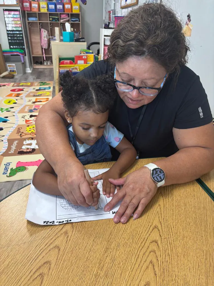 An EduCastle teacher gently guides a toddler&rsquo;s hand as she traces letters on a worksheet