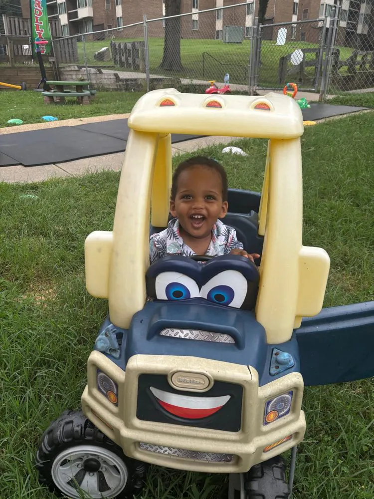 A laughing toddler driving a ride-on toy car on the grass of the EduCastle playground